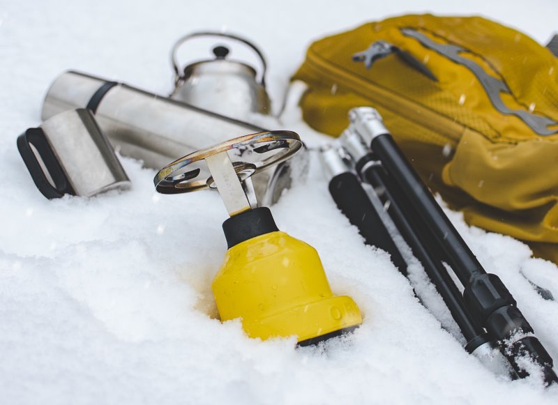 Aufsatz-Gaskocher mit gelber Kartusche liegt im winterlichen Schnee Gelber Kartuschen-Gaskocher liegt neben Outdoor-Equipment im Schnee.
